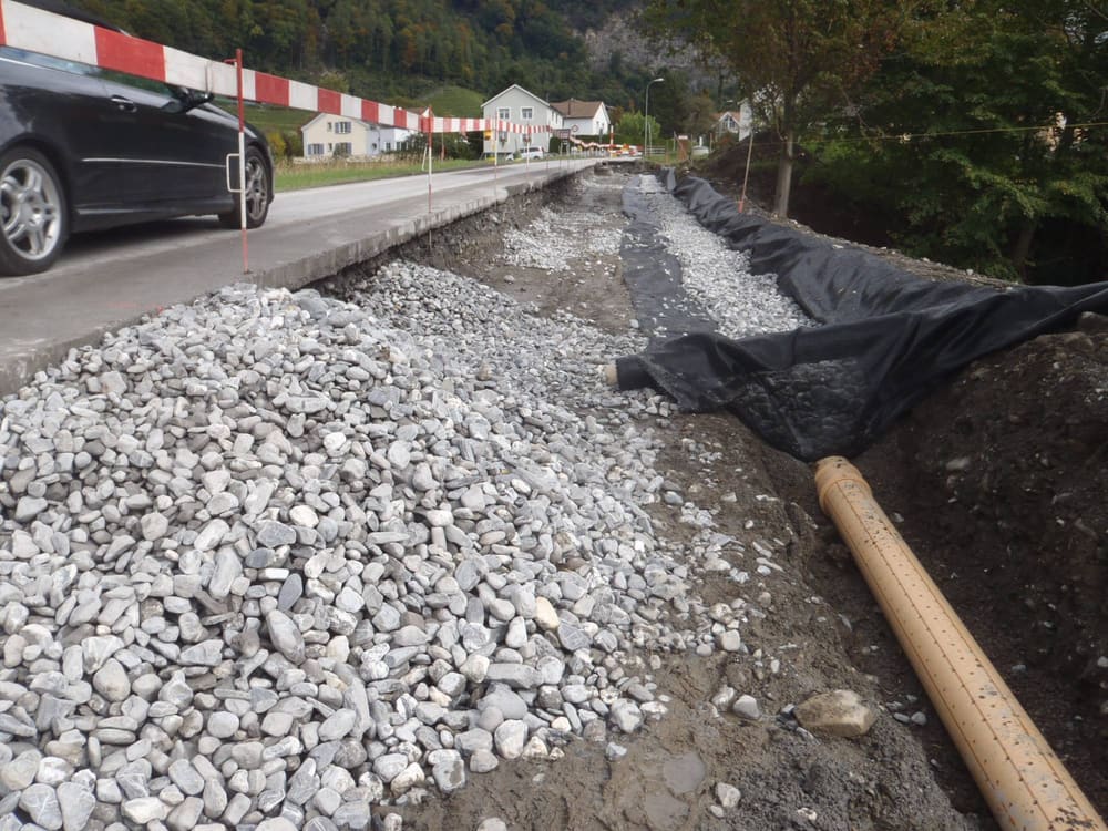 Neue Strassenentwässerung: Sickerleitung mit Filtervlies und Kies neben der bestehenden Fahrbahn, im Hintergrund der Dorfrand von Fläsch