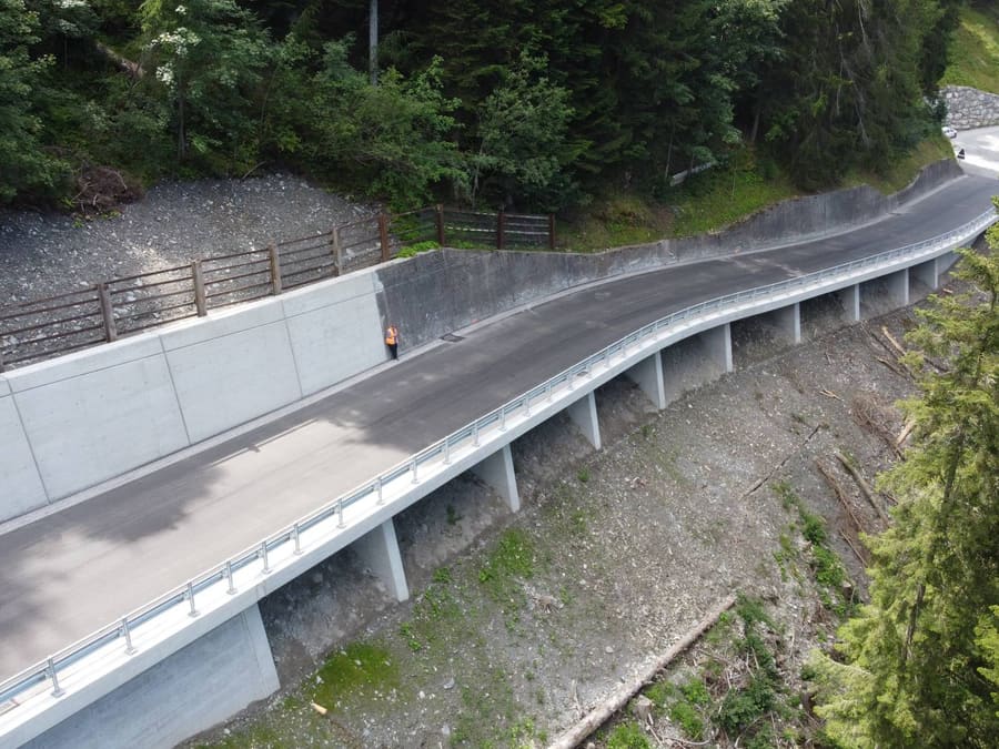Neue Lehnenbrücke Ricaldei aus der Luft: geschwungene Fahrbahn auf schlanken Pfeilern am Waldhang, bergseits eine neue Betonwand mit Holzzaun