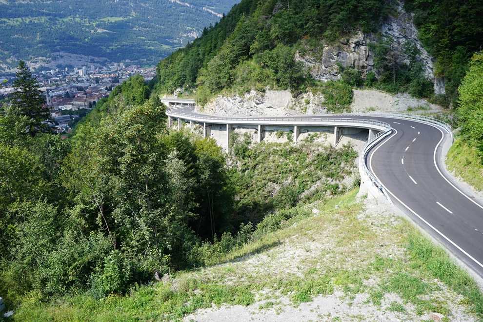 Lehnenbrücke Nasstobel: geschwungene Strassenbrücke auf Stützen entlang einer bewaldeten Felsflanke, frischer Belag mit Leitschranken, im Hintergrund der Talboden mit Siedlung
